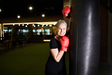 jolly senior woman in sportswear posing with boxing gloves near punching bag and smiling at cameraの写真素材