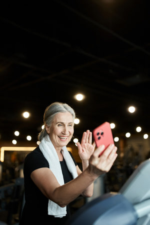 good looking smiling mature woman with towel on shoulders waving at video call while on treadmill