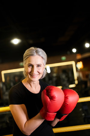 good looking cheerful senior sportswoman with boxing gloves smiling at camera while on ring in gymの写真素材