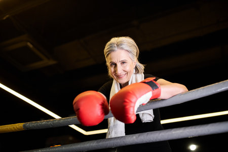 joyous athletic mature woman with towel and boxing gloves exercising on ring and smiling at cameraの写真素材