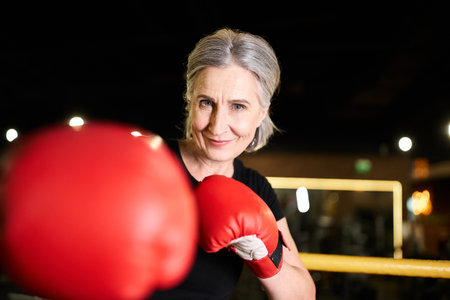 pretty positive senior sportswoman with boxing gloves training on ring and smiling at camera in gymの写真素材