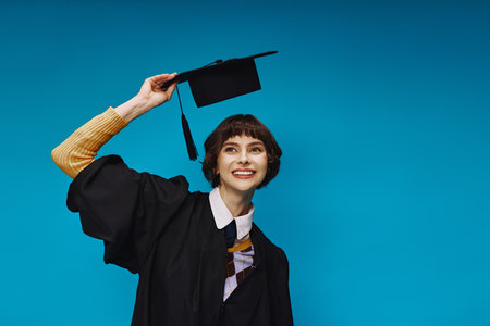 happy college girl wearing black graduation gown holding academic cap on blue background in studioの写真素材