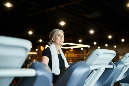 appealing mature sportswoman in black t shirt with towel on shoulders training on treadmill actively