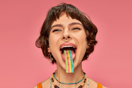 cheerful young woman in her 20s tasting sweet and sour candy strip on her tongue, pink backgroundの写真素材