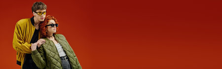 appealing trendy couple in fashionable attires with sunglasses posing on red backdrop, bannerの写真素材