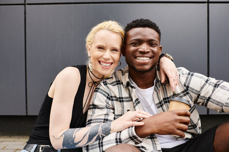 A happy, multicultural couple - boyfriend and girlfriend - sitting next to each other on an urban street near a grey building.の写真素材