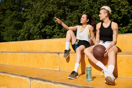 Two young women, athletic friends, sit closely together after playing basketball outdoors on a summer day.の写真素材