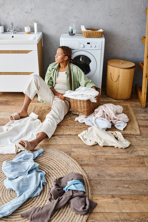 African American woman with afro braids sitting by washing machine in a bathroom, engaged in doing laundry.の写真素材