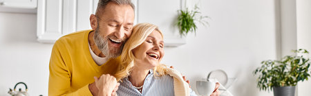 A mature man and woman in homewear share a joyful moment as they laugh together in a cozy kitchen at home.の写真素材