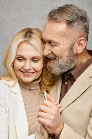 A mature man and woman, dressed elegantly, share a tender hug on a gray backdrop.