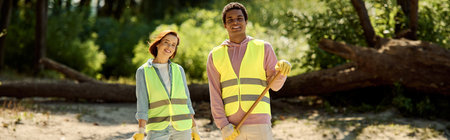 A diverse, socially active couple, in safety vests and gloves, standing together in dirt, cleaning a park with care.の写真素材