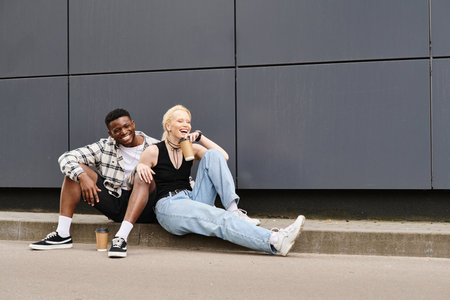 A happy multicultural couple sitting beside each other on the ground near a grey urban buildingの写真素材