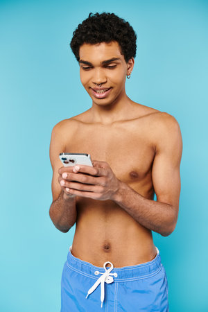 appealing jolly african american man in blue swimming trunks looking at phone on blue backdropの写真素材