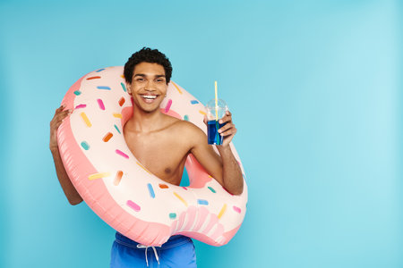 joyous african american man with inflatable donut enjoying refreshing cocktail on blue backdrop