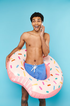 good looking cheerful african american man in swimming trunks with inflatable donut talking by phone