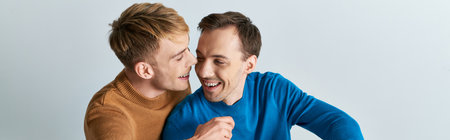Two men, a loving gay couple, in casual attire, standing next to each other on a gray backdrop.