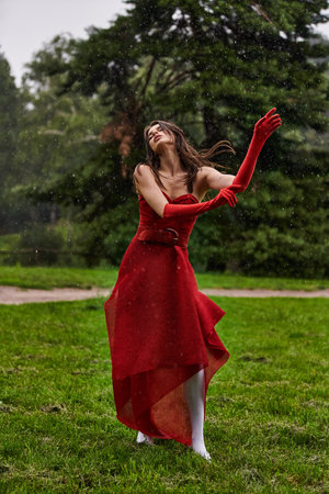 A young woman in a red dress stands gracefully in the rain, enjoying the summer breeze in a natural setting.の写真素材