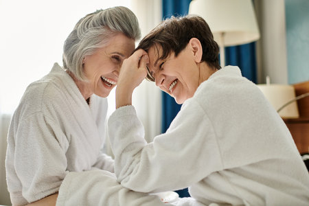 Two senior women share a moment of laughter on a cozy bed.の写真素材