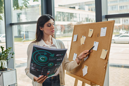 A businesswoman stands in front of board, holding charts in a modern office setting.の写真素材