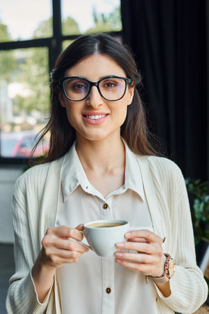 A businesswoman with glasses enjoys a coffee break in a modern office workspace.の写真素材