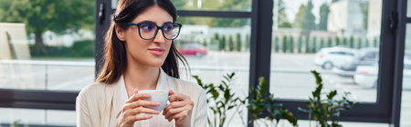 A stylish woman in glasses enjoys a cup of coffee in a modern office, with a hint of focus on productivity and relaxation.の写真素材