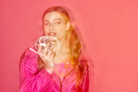 A young woman in a pink dress elegantly holds a mirror ball, creating a dreamy and magical atmosphere in a studio with a pink background.の写真素材