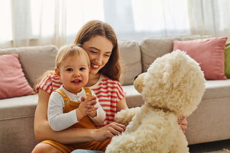 A young mother holding her baby girl lovingly, both smiling, while the baby clutches a teddy bear closely.の写真素材