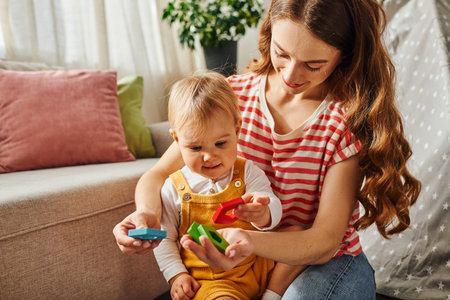 A young mother happily interacts with her toddler daughter, engaging in playful activities on the...