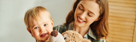 A young mother holds a teddy bear, smiling at her toddler daughter in a heartwarming moment of love...