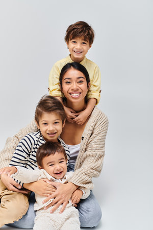 A young Asian mother lovingly poses with her children in a studio, set against a soft grey...