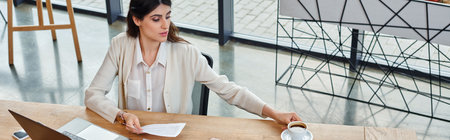A businesswoman focused on work, seated at table with laptop, in modern office setting.