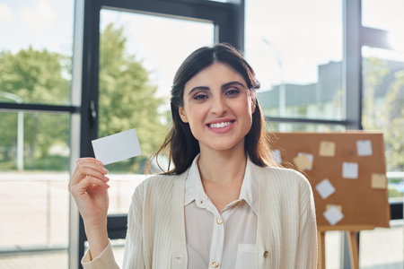 A modern businesswoman holds up a business card in front of a window in a sleek office space.の写真素材