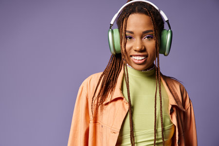 positive african american woman in vibrant attire with wireless headphones on purple backdrop