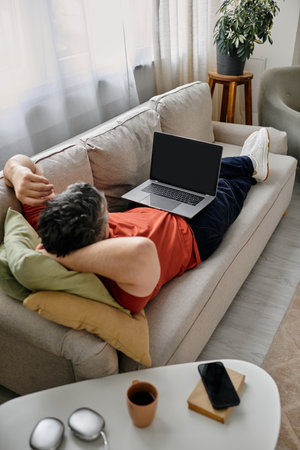 A bearded man in casual attire lies on a white couch with his feet up, working remotely on his laptop.の写真素材