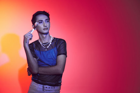 sophisticated androgynous man in stylish attire using makeup brush and posing on vibrant backdropの写真素材