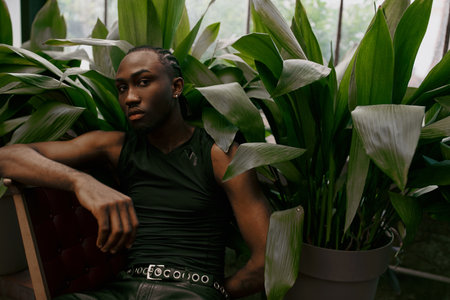 A dapper African American man sits beside a lush potted plant, immersed in nature.