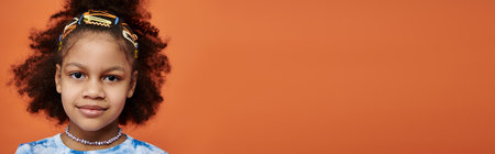 Young Afro-American girl with colorful hair clips, trendy outfit, and orange background, gazes at the camera.の写真素材