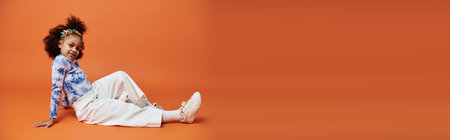 African American girl in a trendy outfit poses against an orange background. She wears white pants, a tie-dye top, and hair clips.の写真素材