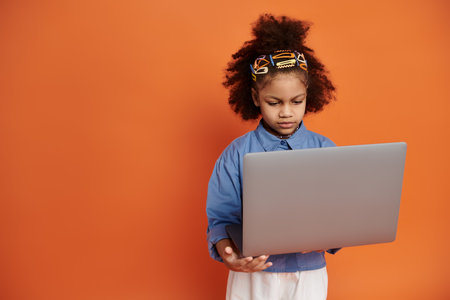 Stylish African American girl in trendy outfit stands in front of orange background, hair in ponytail with clips, holding a laptop.の写真素材