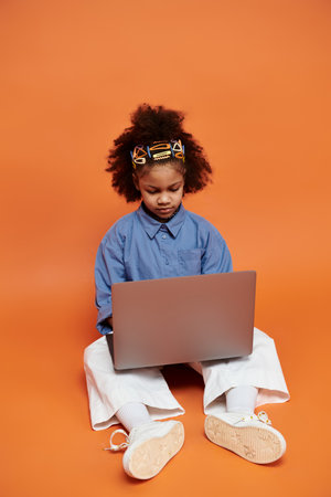 A stylish African American girl with colorful hair clips is sitting on an orange background and...
