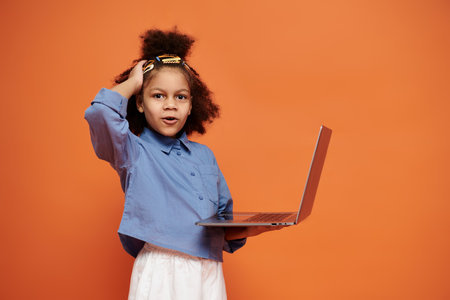 A stylish African American girl with hair clips, trendy outfit, holds a laptop against an orange backdrop.の写真素材