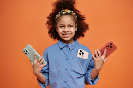 An African American girl in a blue shirt and hair clips stands against an orange background, holding two smartphones and smiling.の写真素材