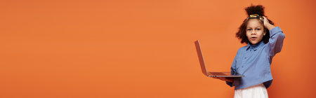 A young African American girl in a trendy outfit holds a laptop in front of an orange background.の写真素材