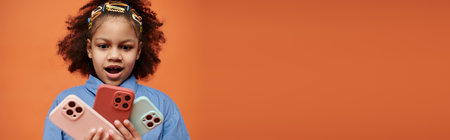 A young African American girl in a blue shirt stares in awe at a collection of colorful phone cases against an orange backdrop.の写真素材