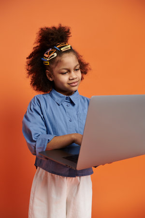 A stylish African American girl with colorful hair clips stands in a trendy outfit, using a laptop against an orange background.の写真素材