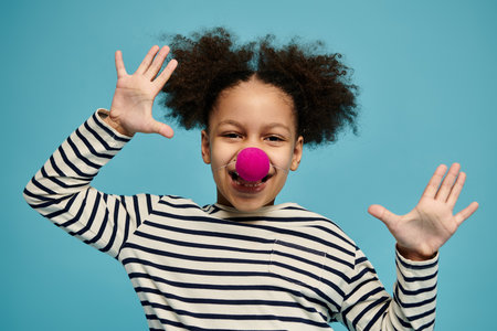 A young African American girl with curly hair smiles cheerfully while wearing a striped shirt and a red clown nose against a blue backdrop.の写真素材