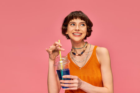 pleased young woman with short brunette hair holding refreshing summer drink on pink backdrop