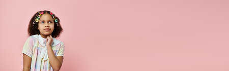 A cute African American girl with colorful hair clips stands against a pink background, thoughtfully gazing upwards.の写真素材