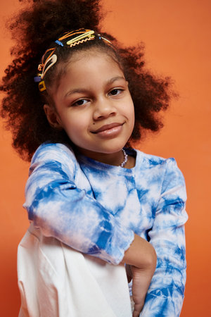 A young African American girl wearing a trendy outfit with her hair styled with colorful clips stands against a vibrant orange background.の写真素材