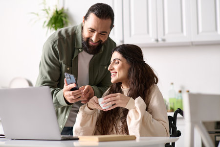 appealing loving couple of bearded man and disabled woman looking at phone in kitchen at homeの写真素材